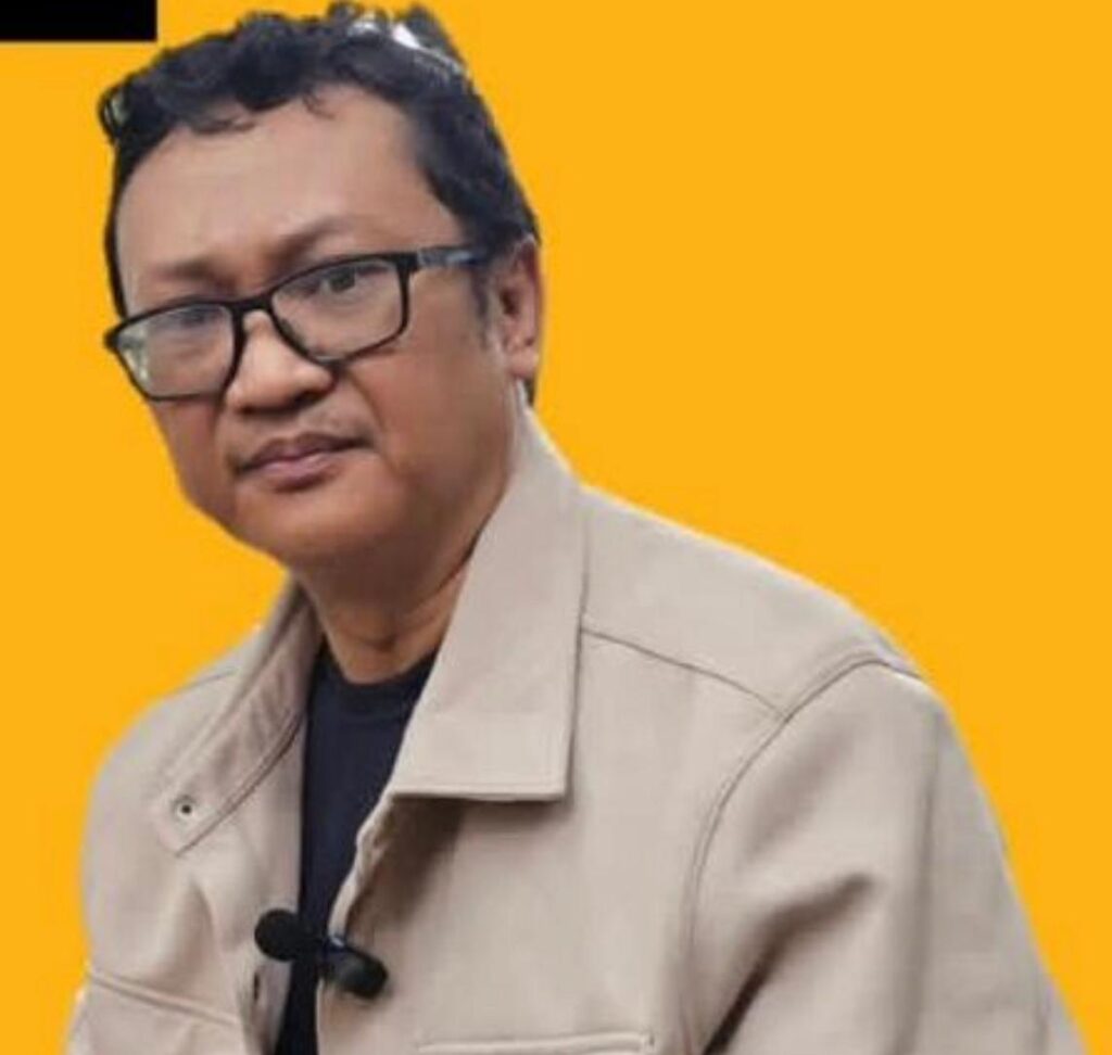 Whatsapp image 20260701 at 14 20 29 catrawarta Portrait of a man wearing glasses and a beige jacket with a clip on microphone against an orange background